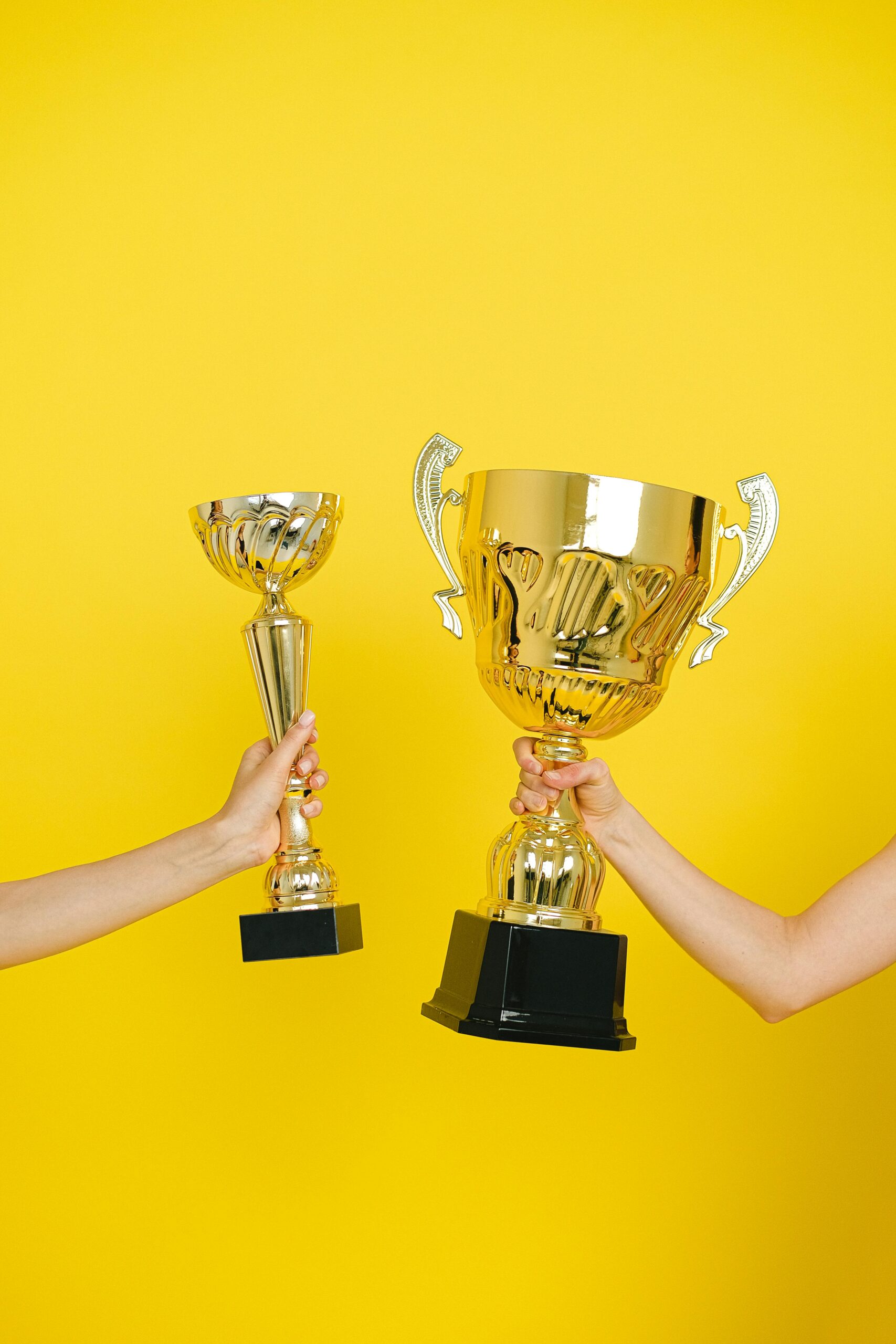 Two hands holding trophies against a vivid yellow backdrop, symbolizing success.
